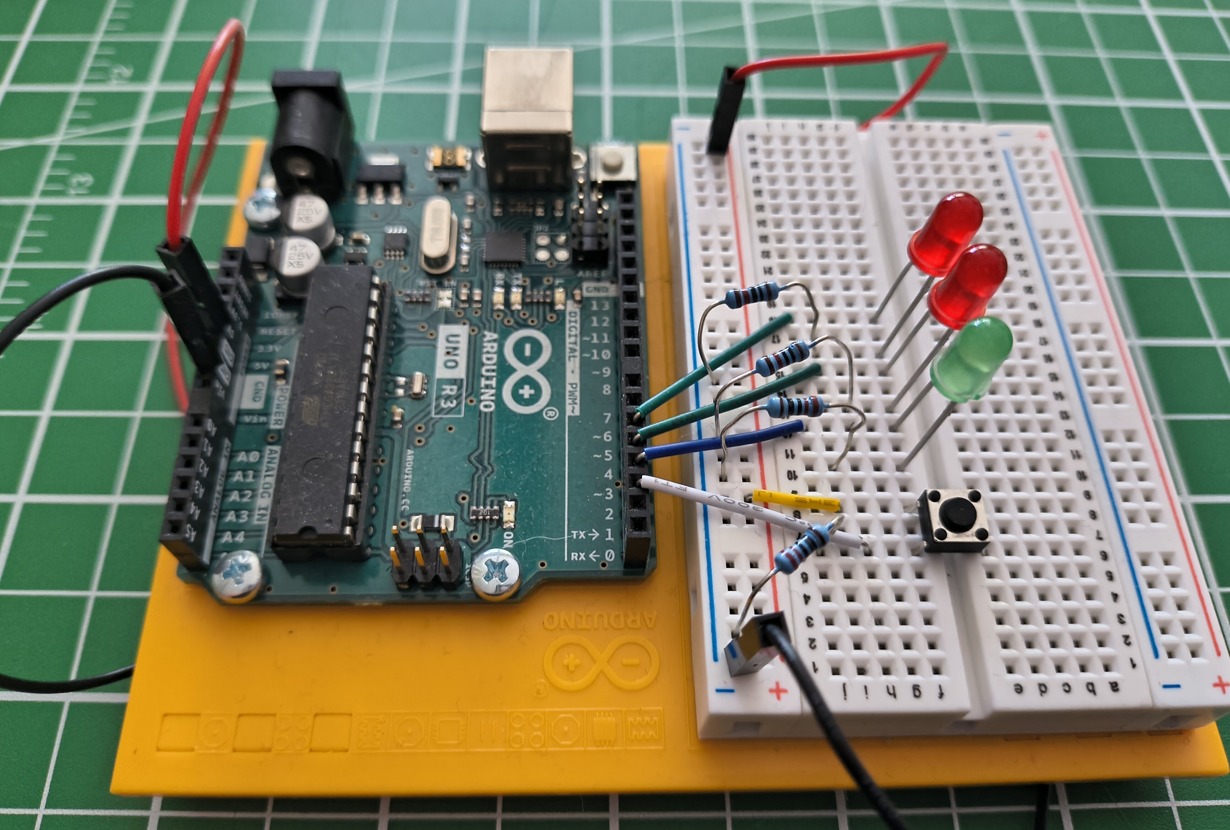 An Arduino Uno on a yellow base wired to a breadboard holding three LEDs, four resistors, and a pushbutton, connected by coloured jumper wires.