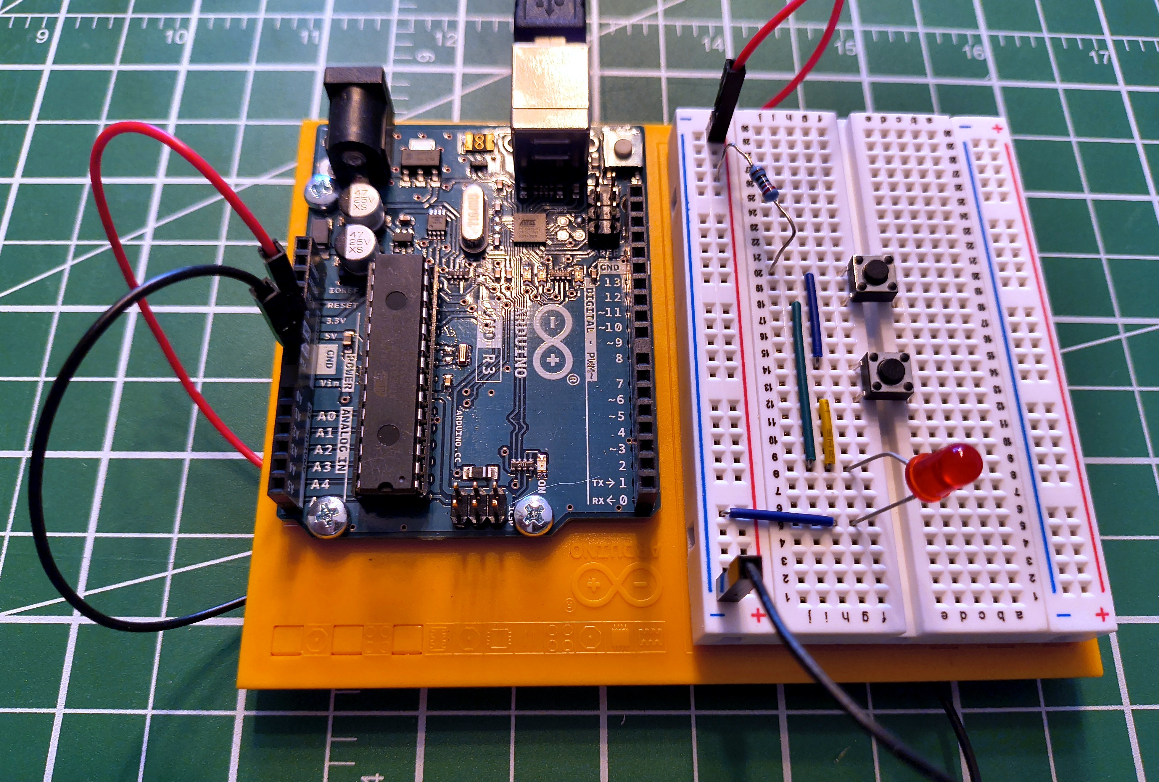 An Arduino Uno connected to a half-size breadboard with two tactile buttons, a resistor, and an LED wired in parallel, showing power rails, jumper wires, and component placement.