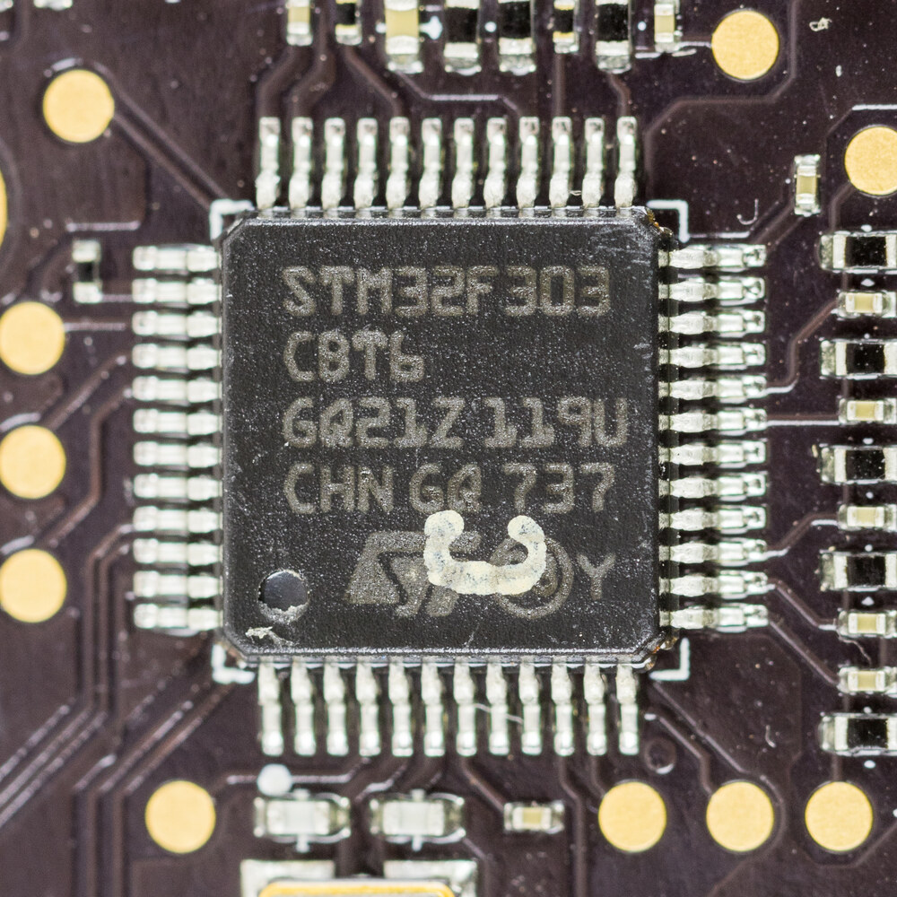 An extreme macro photo of an STM32F303 microcontroller soldered onto a circuit board, showing its flat rectangular body with fine metal legs on all four sides.