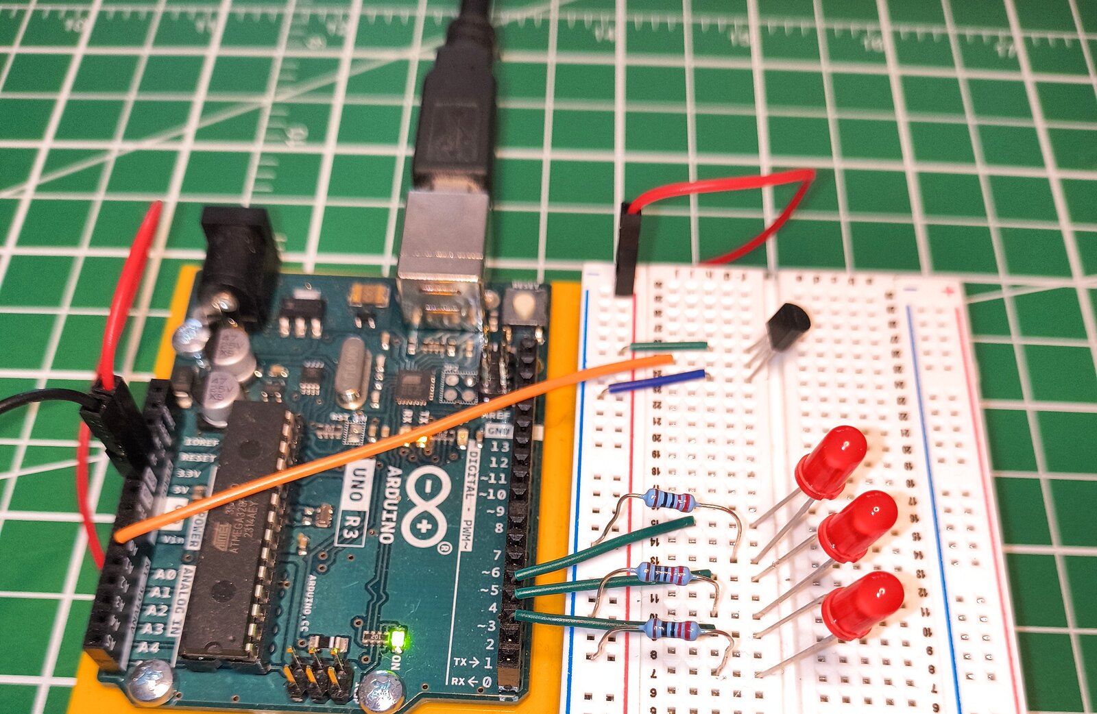 An Arduino Uno on a breadboard wired to a black TO-92-packaged MCP9700A temperature sensor, connected by jumper wires to the 5V rail, ground rail, and analog pin A0. Three LEDs are also visible on the board, wired but not yet used.